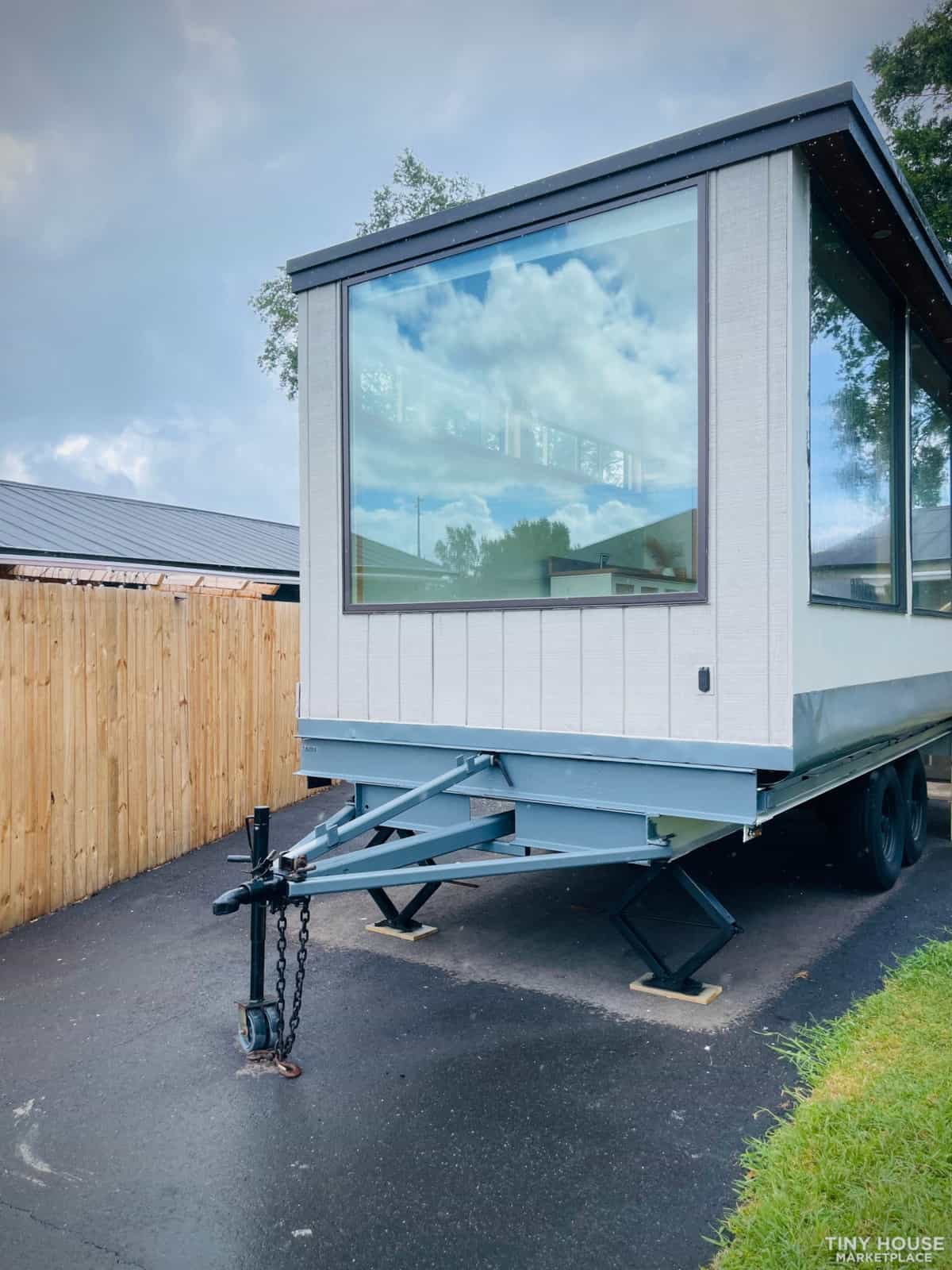 Front exterior showing massive picture window, white board-and-batten siding, and teal dual-axle trailer with hitch