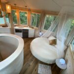 Charming treehouse interior featuring a round moon-shaped bed with a canopy, surrounded by large win.