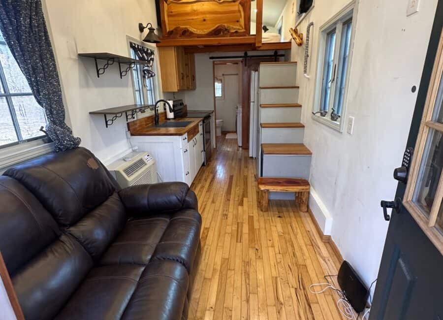 Compact tiny house living space with a loft, wooden stairs, and large windows overlooking a creek.