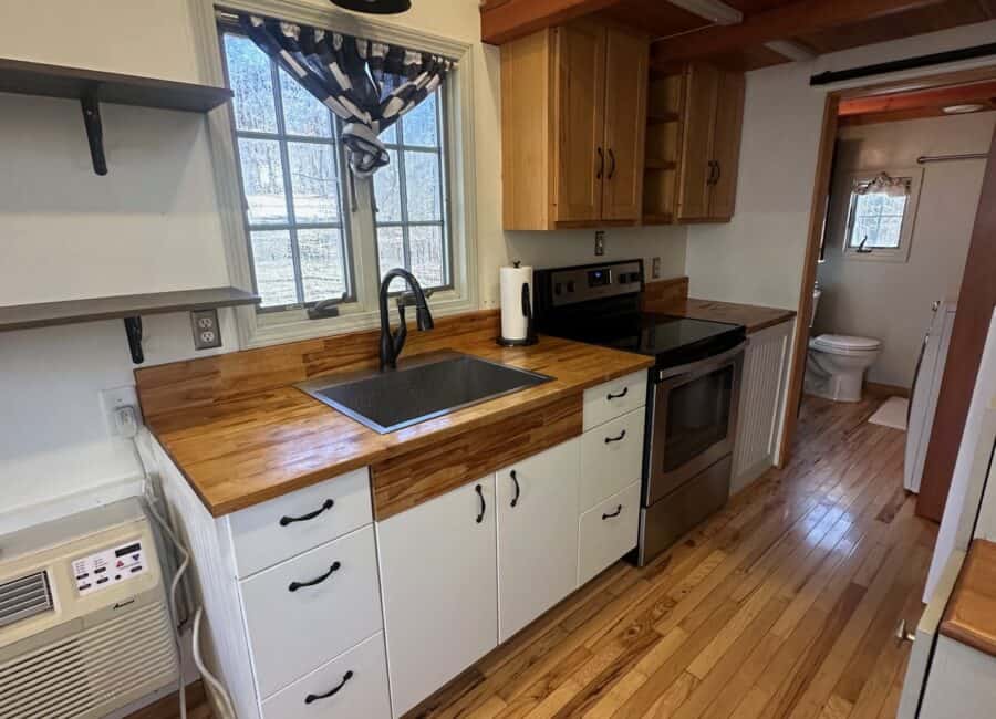 Compact tiny house kitchen with wooden countertops and large window overlooking nature.