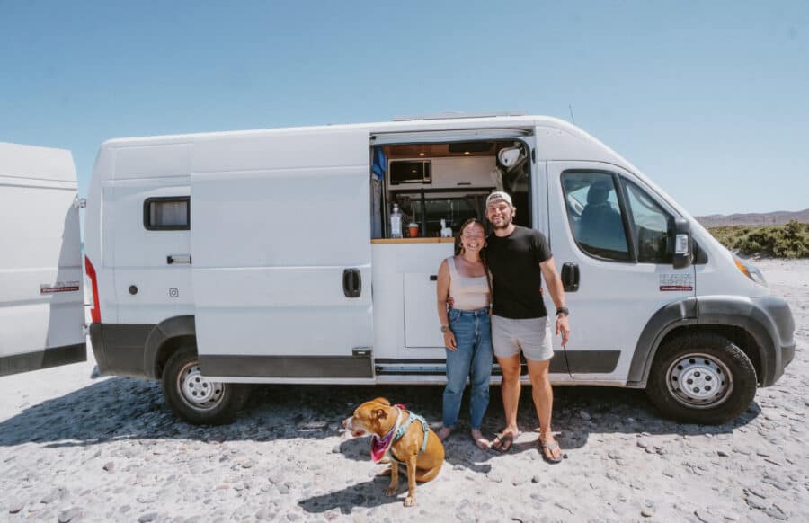 Happy couple with dog in front of a white tiny house van on a sunny day.