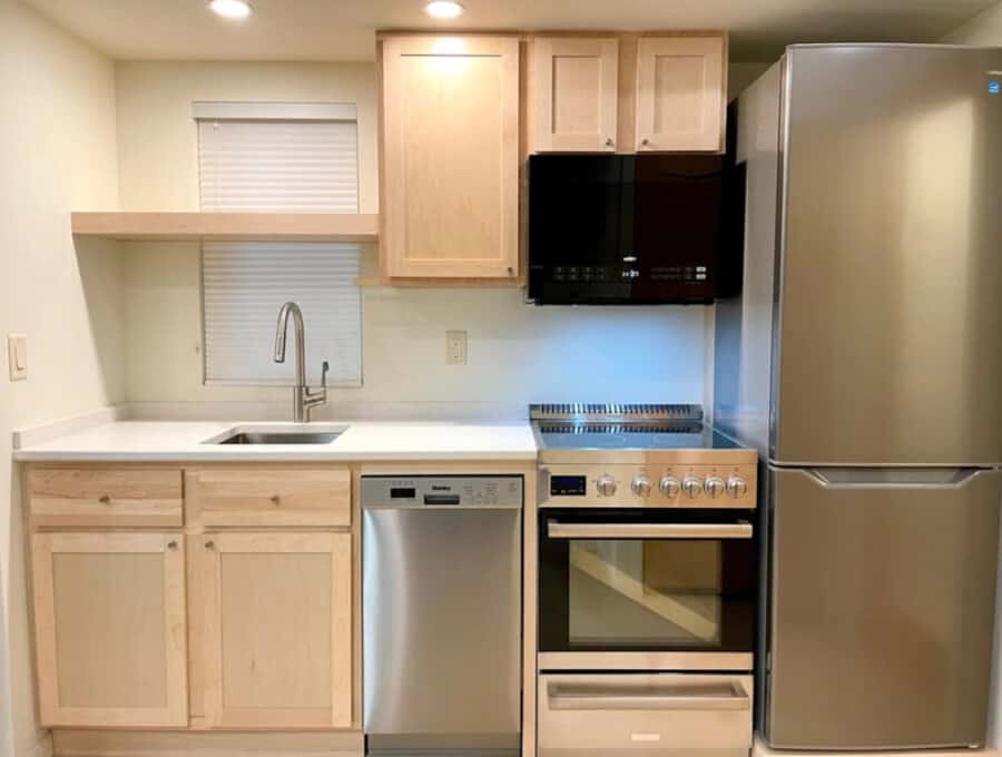 Compact modern tiny house kitchen with stainless steel appliances and light wood cabinets.