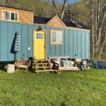 Whimsical farmhouse tiny house on wheels with vibrant blue exterior and cozy outdoor seating area.