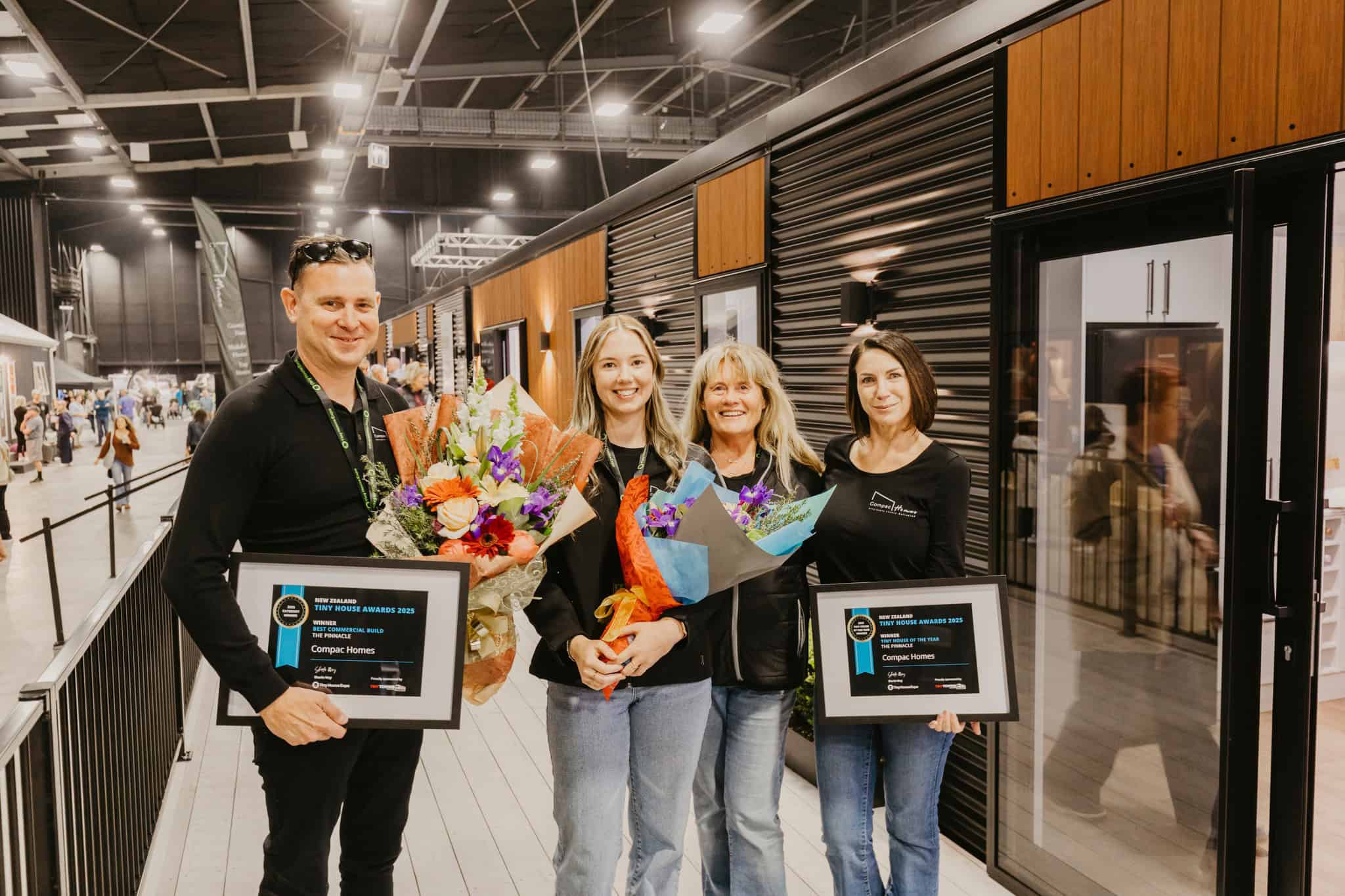 Four members of a tiny house building team holding framed New Zealand Tiny House Awards certificates and flowers
