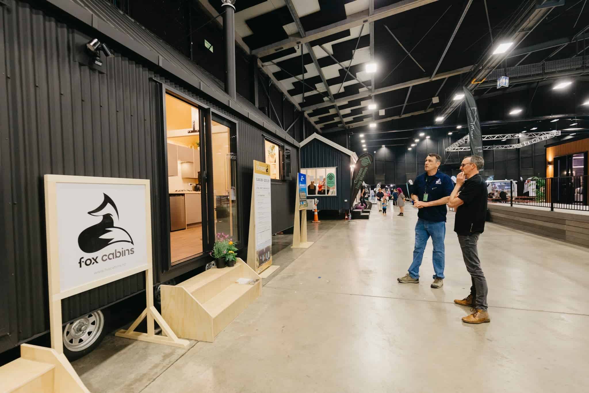Row of tiny homes and cabins displayed inside a large indoor pavilion with visitors browsing
