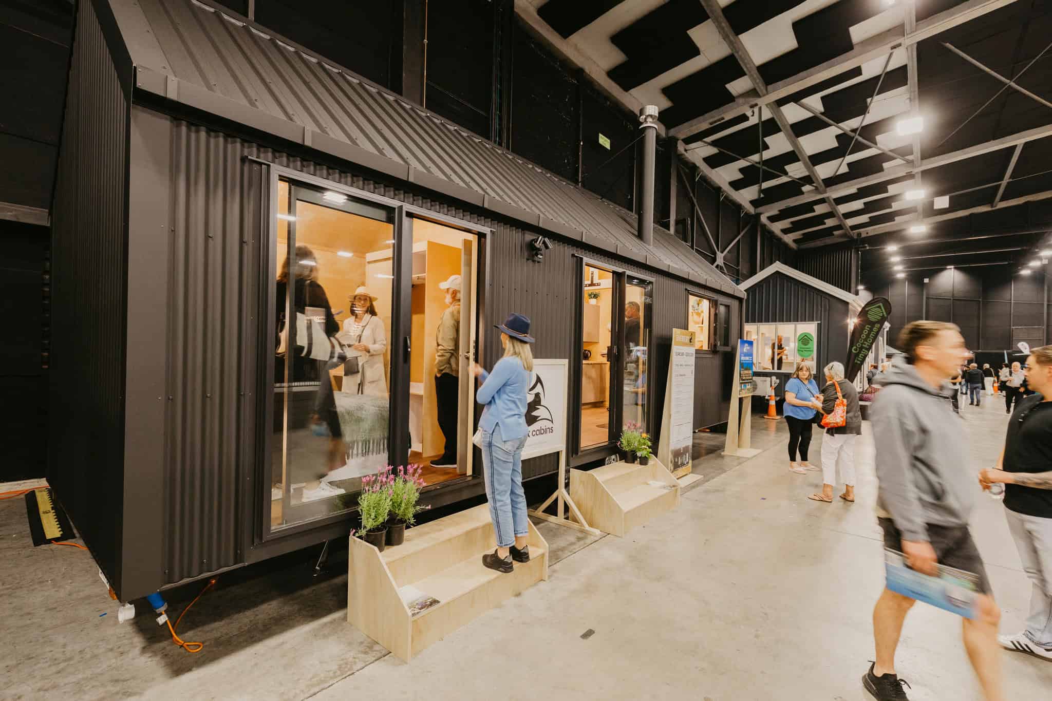 Visitors stepping into a black-clad tiny house on wheels inside the Tiny House Expo pavilion at Mystery Creek
