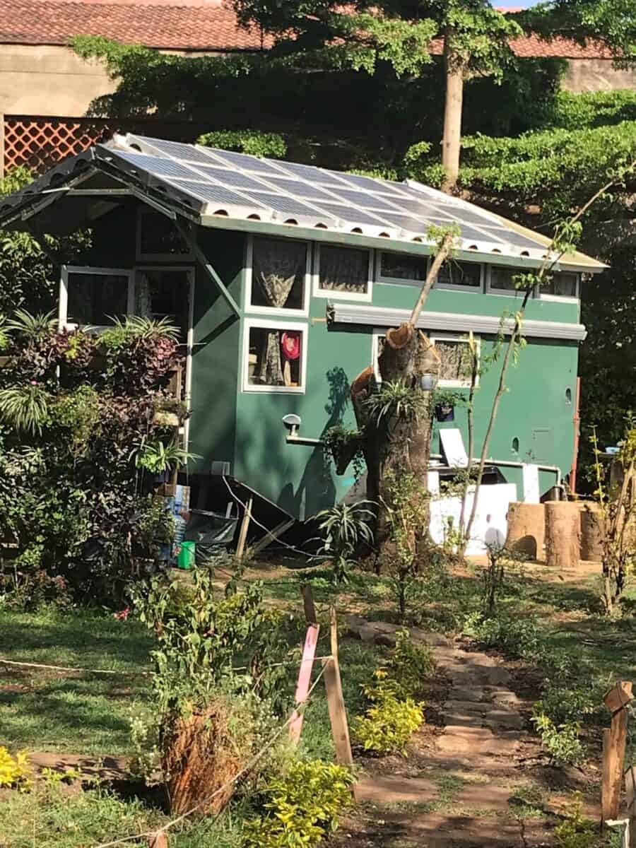 Small green tiny house with solar panels and standing loft in a lush garden setting.