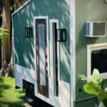 Compact tiny house with green siding, large windows, and air conditioning unit on the side.