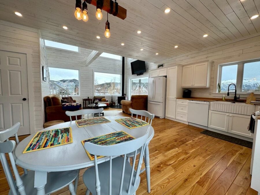 Bright interior of a tiny cabin with white walls, wooden floors, and large windows offering scenic v.