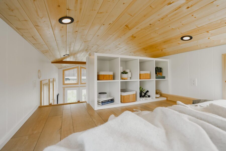 Compact tiny house interior with wooden ceiling and white shelving unit.