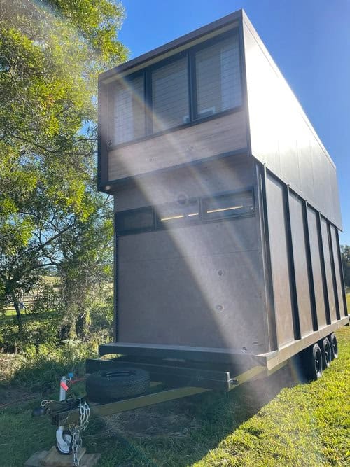 Two-story tiny house on wheels with sleek black exterior and large windows, set outdoors under a cle.