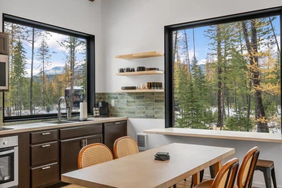 Bright off-grid cabin kitchen featuring large windows overlooking lush forest landscape.