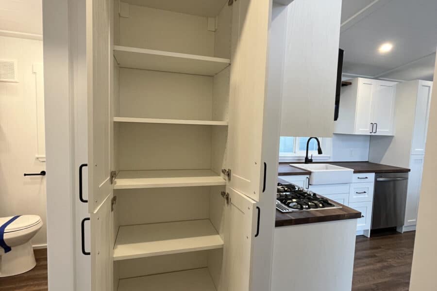 Built-in white storage cabinet inside a tiny house with kitchen and bathroom visible.