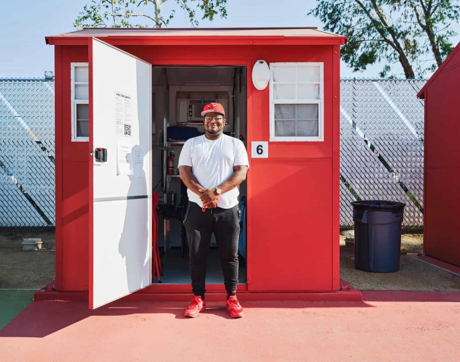 A resident standing in the open doorway of a red 64-square-foot Pallet tiny home showing the air conditioner, shelving and fire extinguisher inside