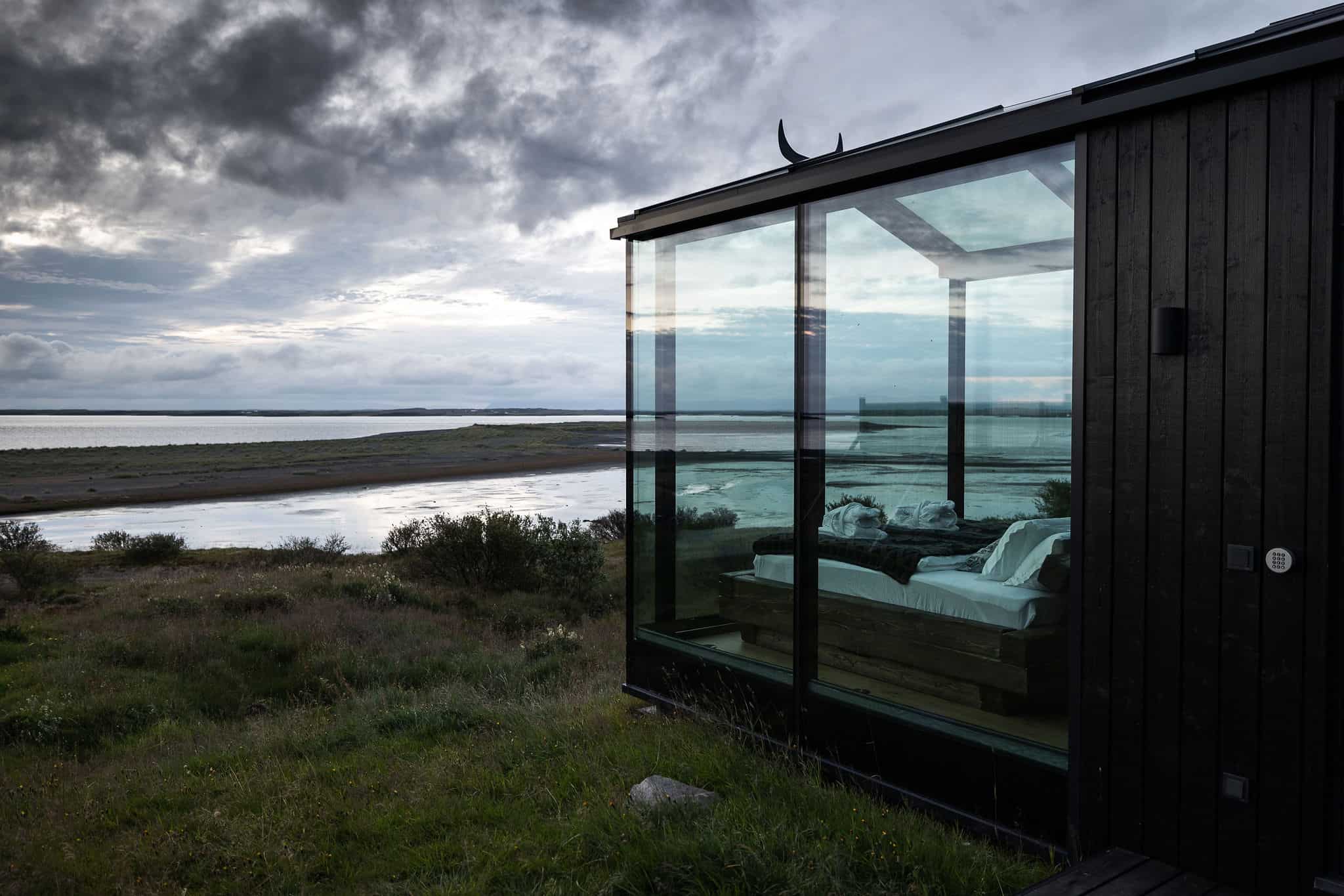 Side view of a Panorama Glass Lodge showing the bed through the glass wall above a river