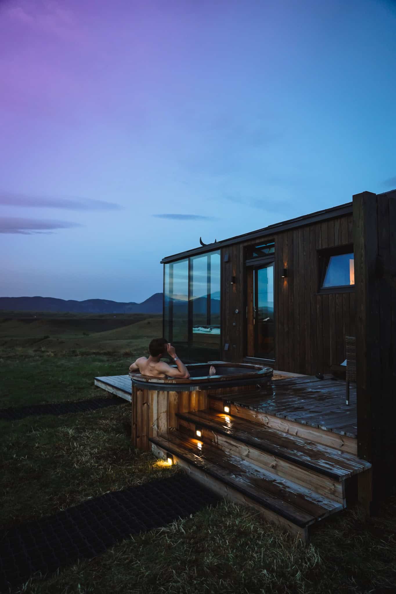 Person relaxing in a private hot tub beside a Panorama Glass Lodge at dusk