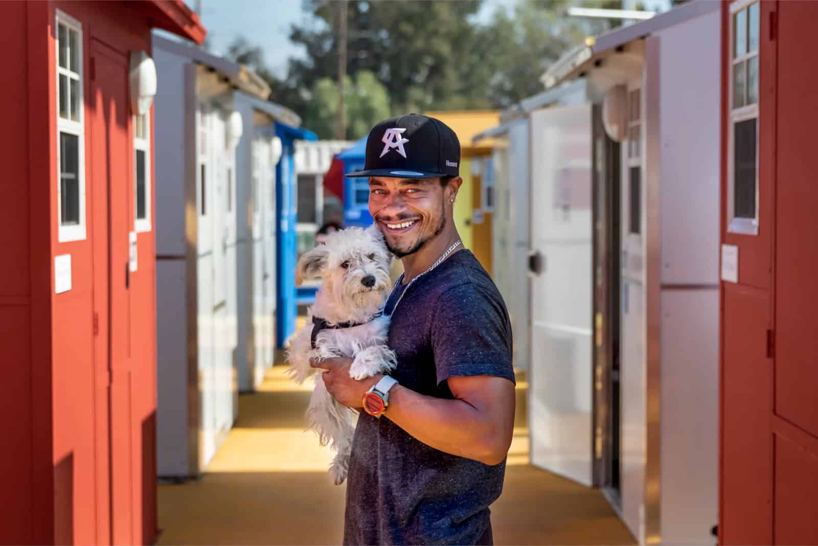 A smiling resident holding a small white dog in the pathway between red and blue tiny homes at a Los Angeles village