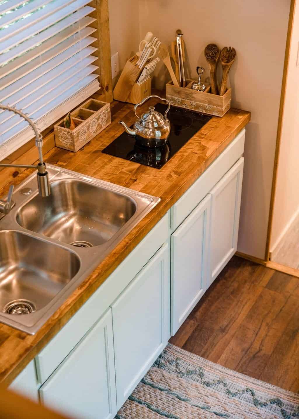 Prana tiny house kitchen with a butcher-block counter, double stainless sink, gooseneck faucet, flush two-burner cooktop, and sage-blue cabinets