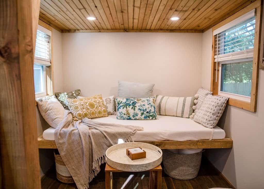 The Prana tiny house living nook, a cushioned built-in daybed with pillows beneath a pine ceiling and windows on two sides