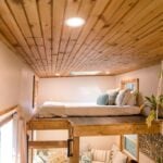 Sleeping loft in the Prana tiny house with a skylight and pine ceiling, overlooking the cushioned living nook below