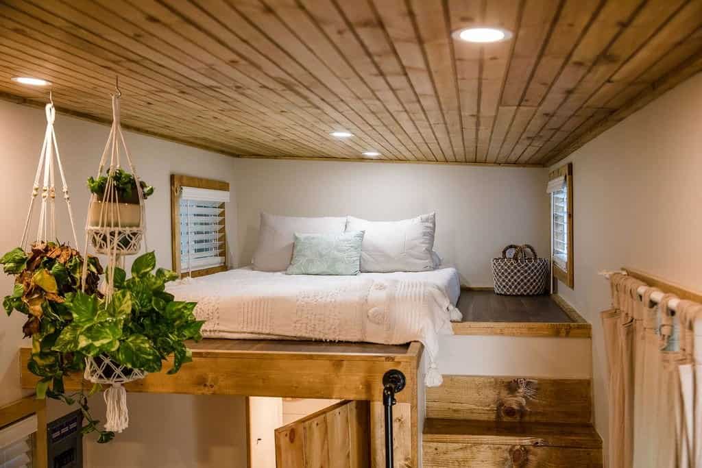 Master loft in the Prana tiny house with a queen bed, white bedding, hanging plants, windows, and open stairs