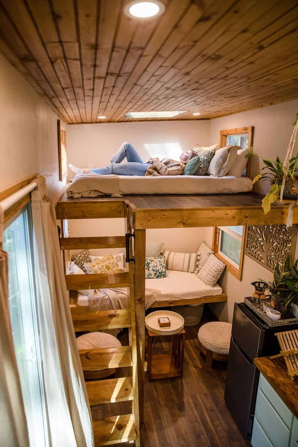 Interior of the Prana tiny house showing a second loft above a pillow-filled living nook, an open staircase, mini-fridge, and the glass entry door