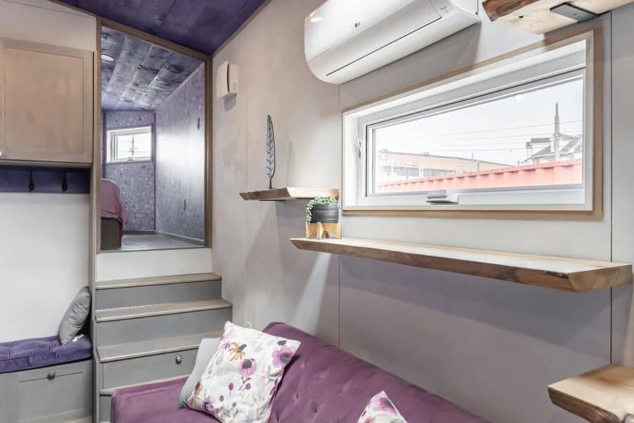 Interior view of a tiny house with a purple sofa, wooden shelves, and large window.