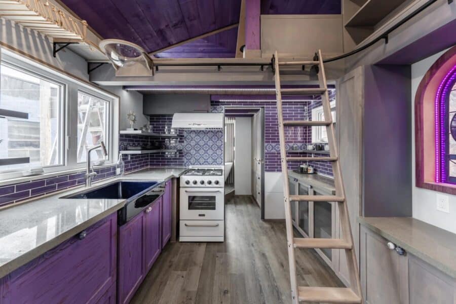 Compact tiny house kitchen with purple cabinets and loft ladder.