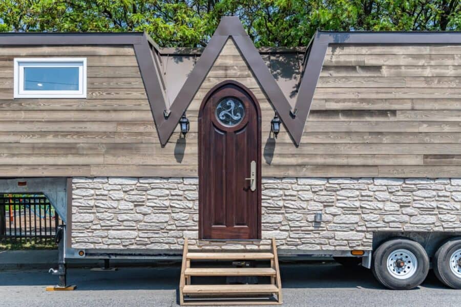 Small tiny house with W-shaped roof and wooden door, on a trailer.