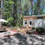 Rasa tiny home with white board-and-batten and cedar siding on a wooded deck with a picnic table and fire pit