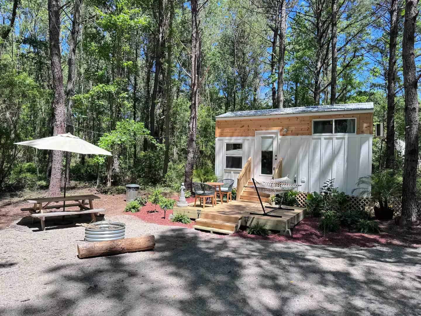 Rasa tiny home with white board-and-batten and cedar siding on a wooded deck with a picnic table and fire pit