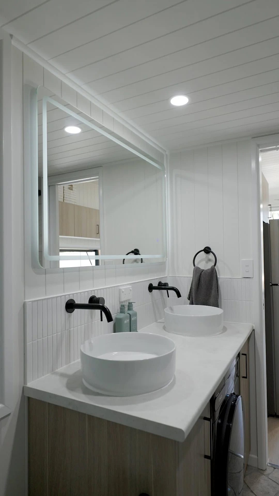 A full double vanity in the Burleigh 9.6 with two round white vessel basins, twin black wall-mounted taps, a backlit mirror, and a built-in washing machine alongside