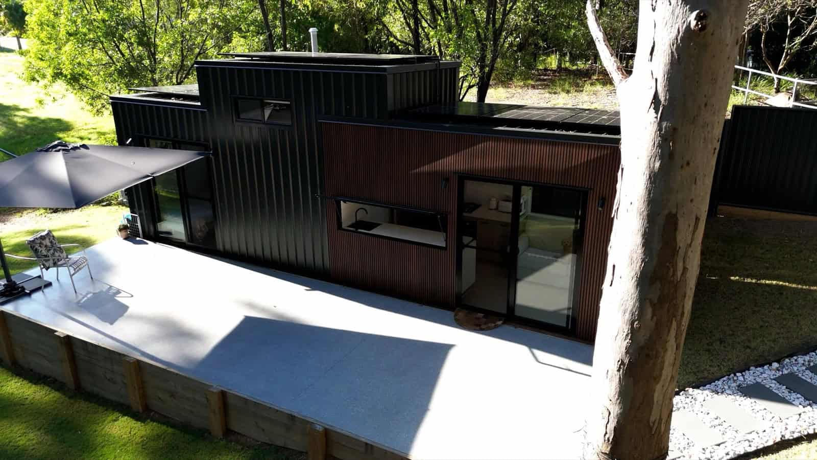 The Burleigh 9.6 by Removed Tiny Homes, a custom tiny house with an angular stepped roofline and wraparound NewTechWood timber-look cladding, set on a concrete deck among gum trees