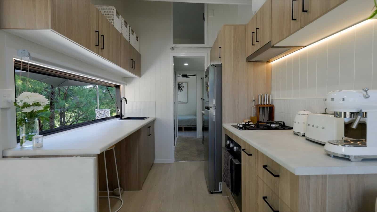 The Japandi-style galley kitchen of the Burleigh 9.6 with light timber cabinetry, white stone benchtops, black hardware, a window over the sink, and SMEG appliances