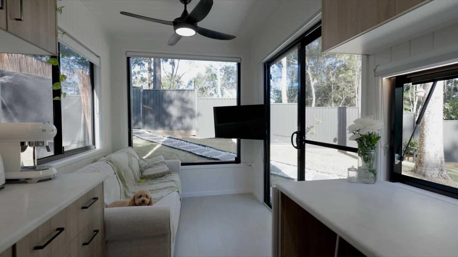 The Burleigh 9.6 living area with a sofa, a wall-mounted swivel TV, large picture windows and a sliding glass door framing the bush, and a ceiling fan