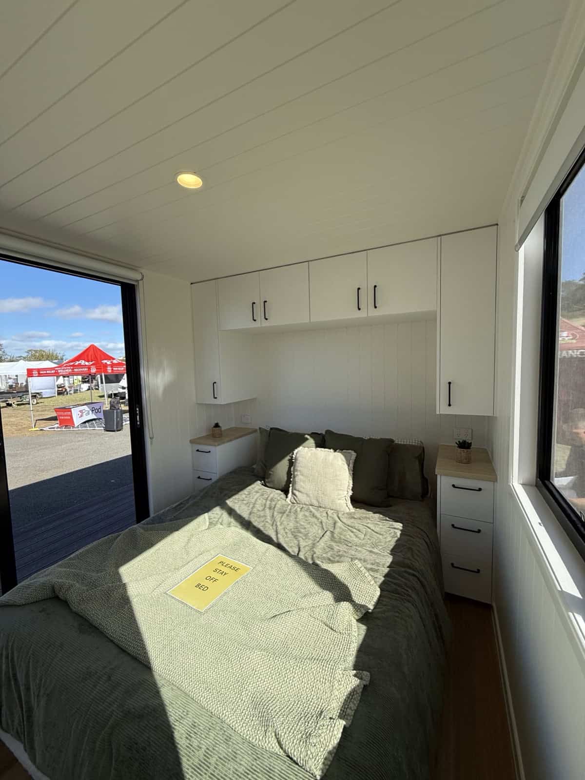 The full-size downstairs bedroom in the Cabarita tiny house with a queen bed, overhead cabinetry, built-in bedside tables, and windows on both sides