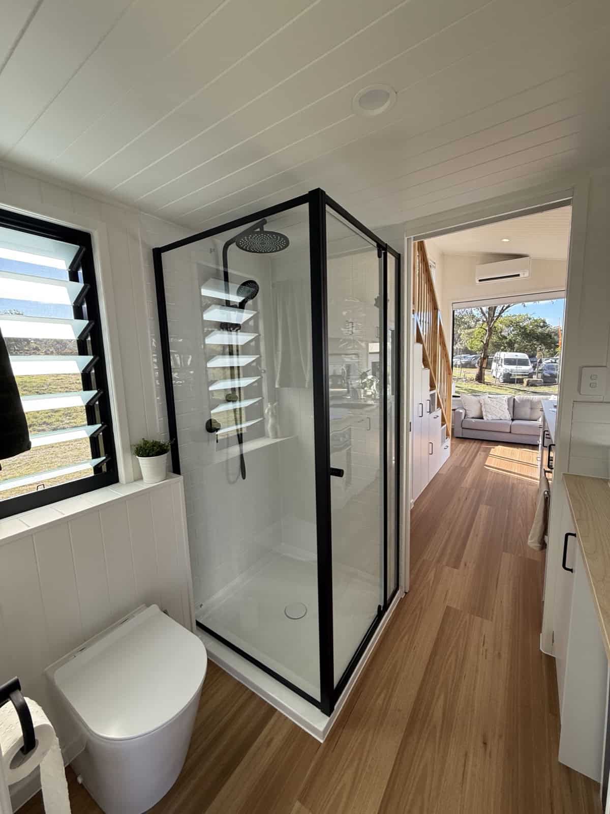 The Cabarita walk-through ensuite with a black-framed glass shower, rainfall head, tiled niches, a modern toilet, and a louvre window