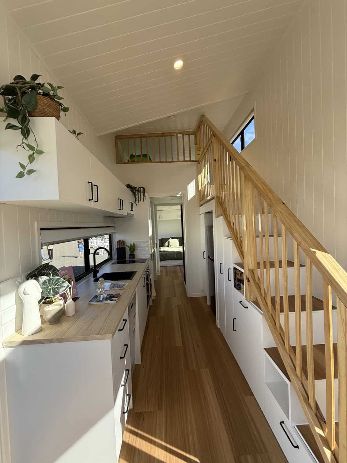 Interior of the Cabarita tiny house looking down the length, with a timber kitchen on the left, full staircase to the loft on the right, and the bedroom visible at the far end