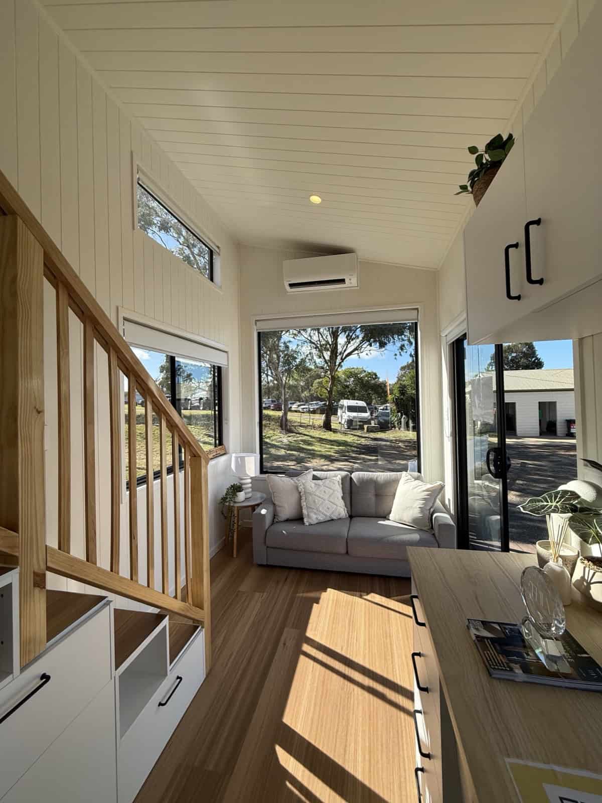 The Cabarita living area with a grey sofa beneath a tall signature picture window, clerestory glazing above, and a sliding glass door to the outside