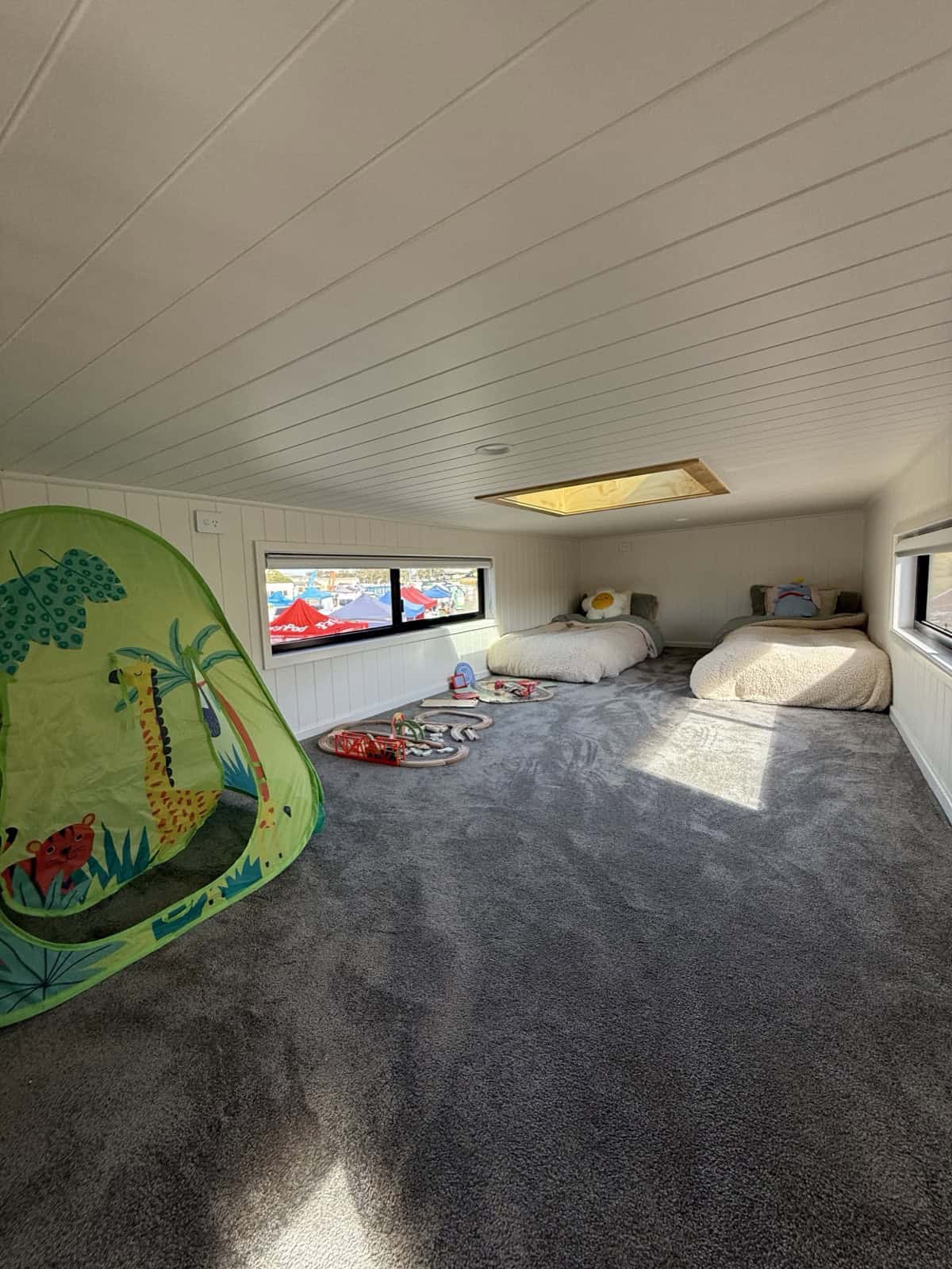 The spacious carpeted upstairs loft in the Cabarita tiny house set up with two beds, a skylight, and windows, with room for a play area