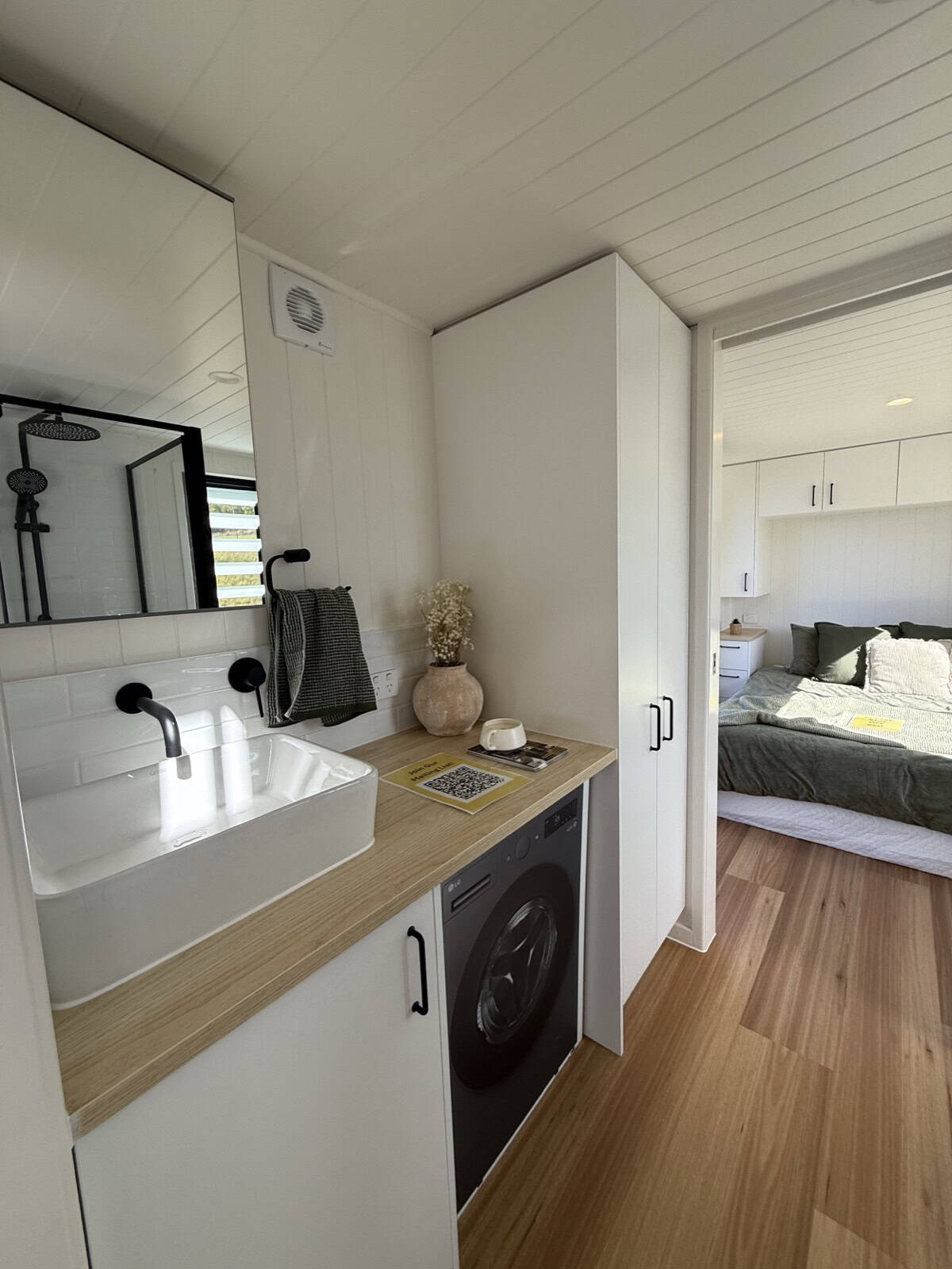 The Cabarita ensuite vanity with a white vessel sink, black tap, large mirror, and a front-loading washing machine built in beneath the bench, with the bedroom visible beyond
