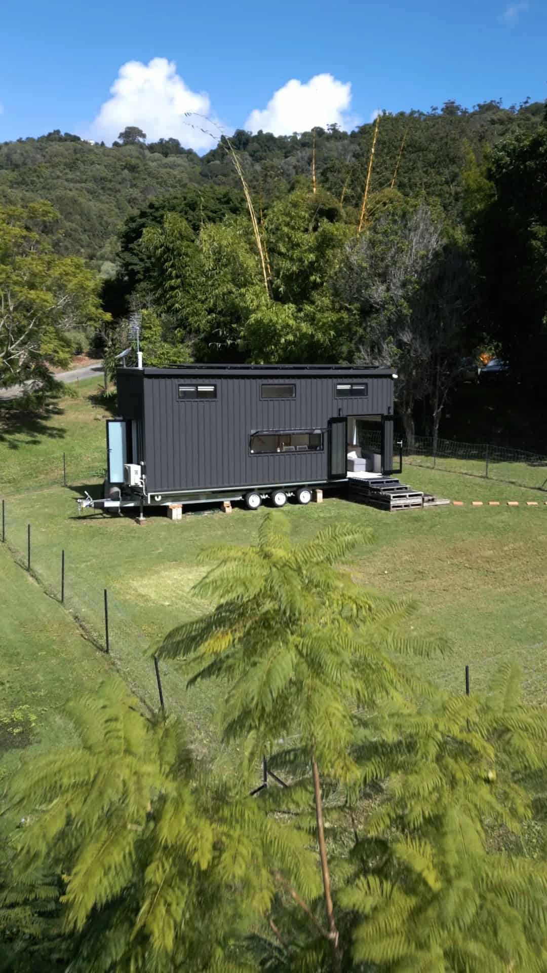 The Coolangatta 8.4 by Removed Tiny Homes, a custom tiny house in dark monument cladding on a trailer, set in a green valley with forested hills behind