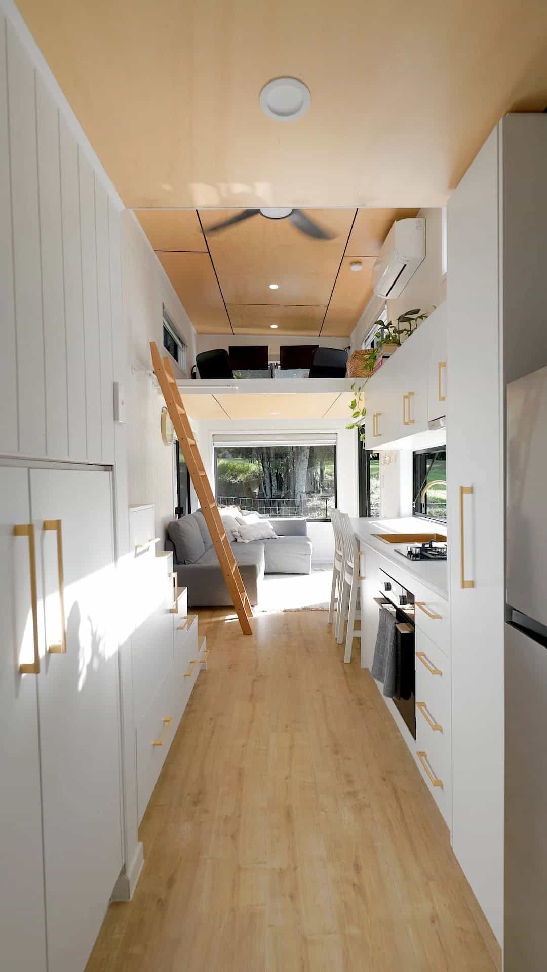 The Coolangatta interior with a custom storage staircase, a floating workspace loft above the living area, a grey sofa beneath a picture window, and a white galley kitchen