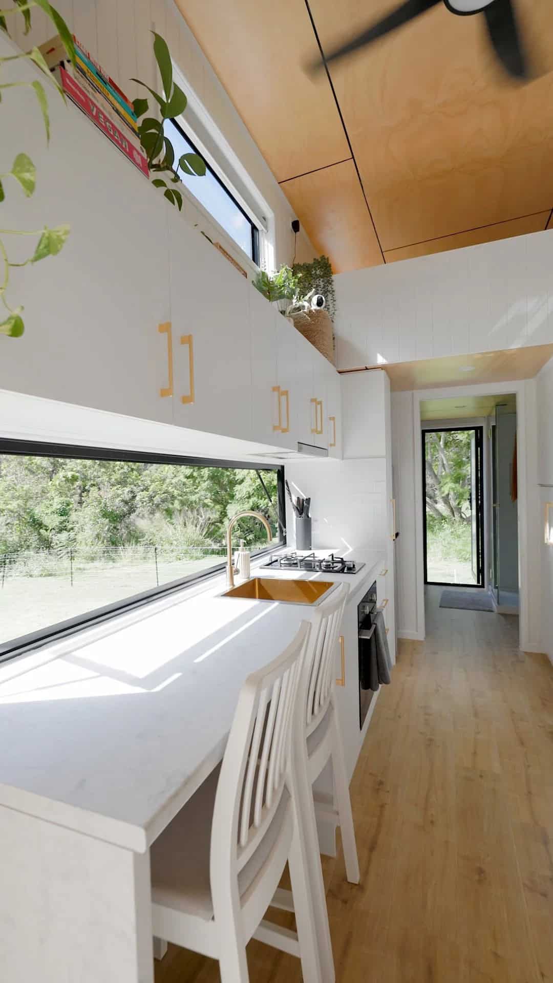 The Coolangatta kitchen with a white stone benchtop extending to a window-side dining bench, brass tap and sink, gas cooktop, and a long picture window framing the outdoors