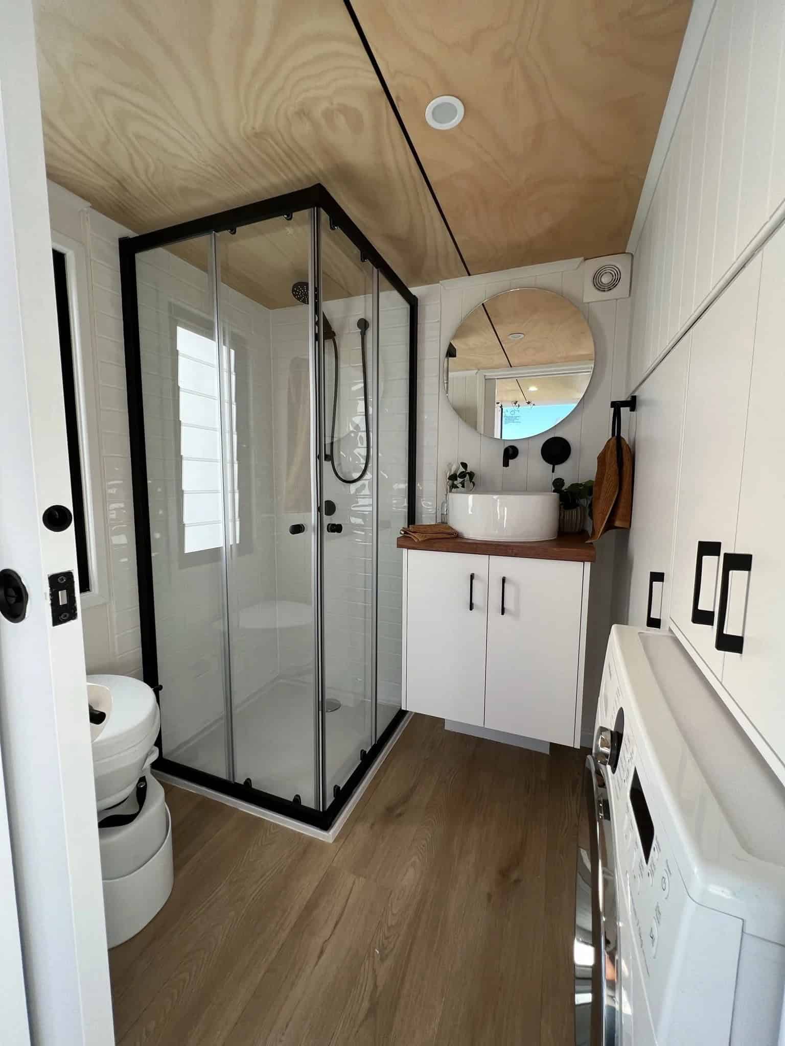 The Currumbin bathroom with a black-framed glass shower, a composting-style toilet, a timber vanity with a white vessel sink and round mirror, and a built-in washing machine