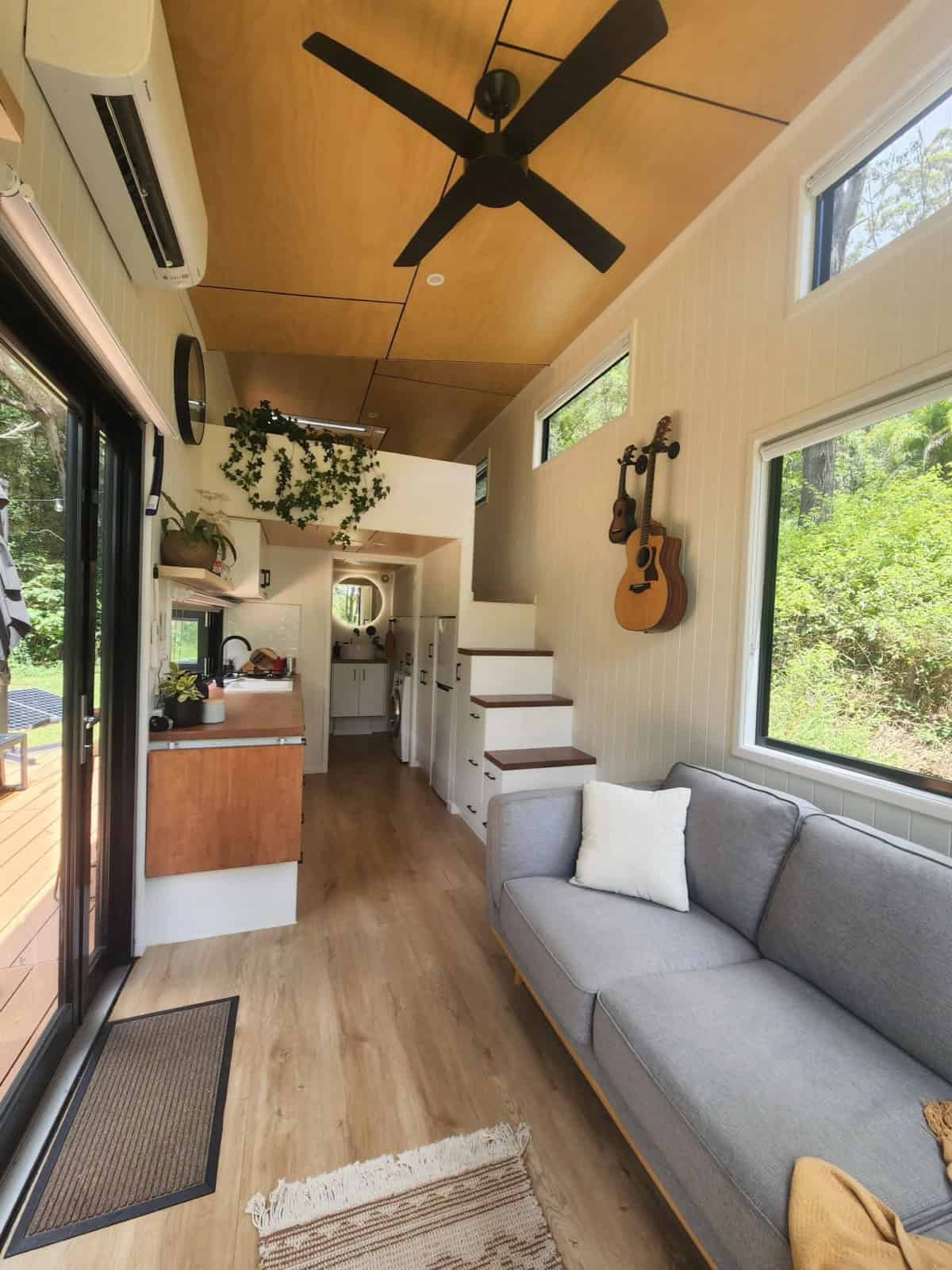 Interior of the Currumbin looking toward the kitchen, with a grey sofa beneath a large picture window, a timber storage staircase to the loft, and a vaulted timber ceiling
