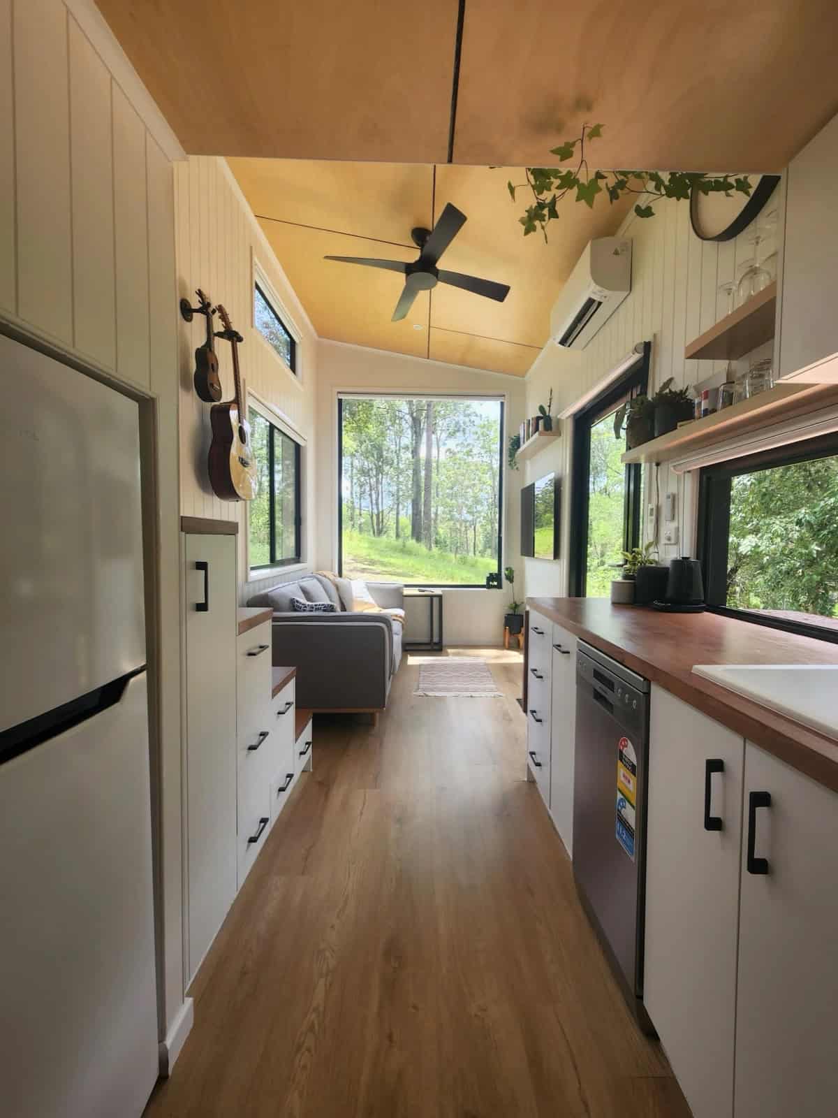 The galley kitchen of the Currumbin looking toward the living area, with a dishwasher, timber benchtops, open shelves, and a picture window framing forest views