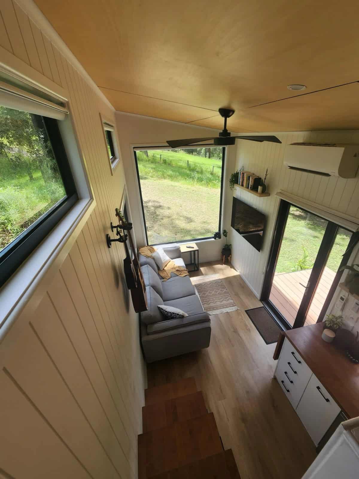 The Currumbin living area seen from the staircase, with a grey sofa beneath a large picture window, a ceiling fan, and a sliding glass door to the deck