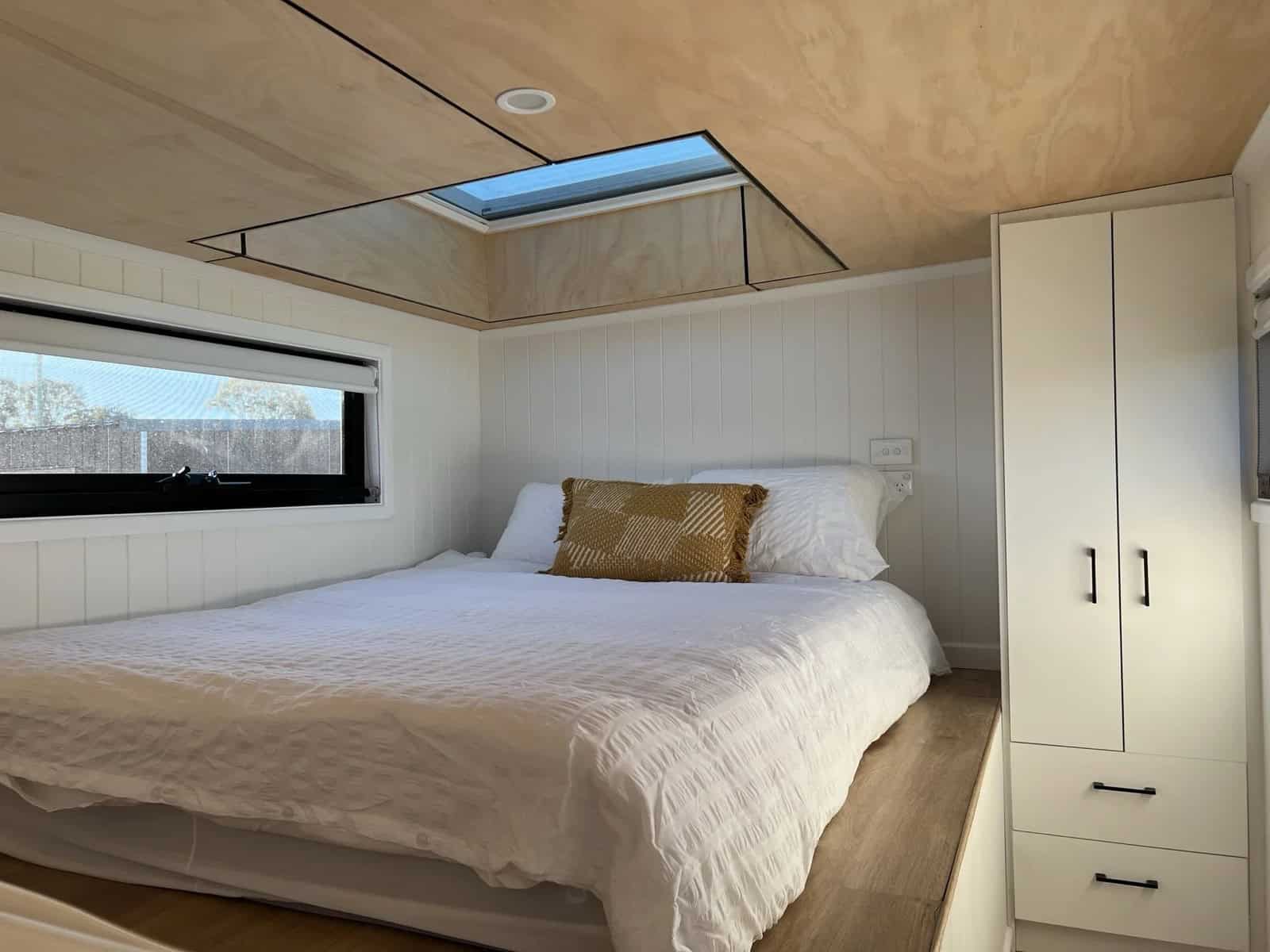 The Currumbin loft bedroom with a queen bed beneath a large skylight, a window alongside, and a full-height white wardrobe with drawers