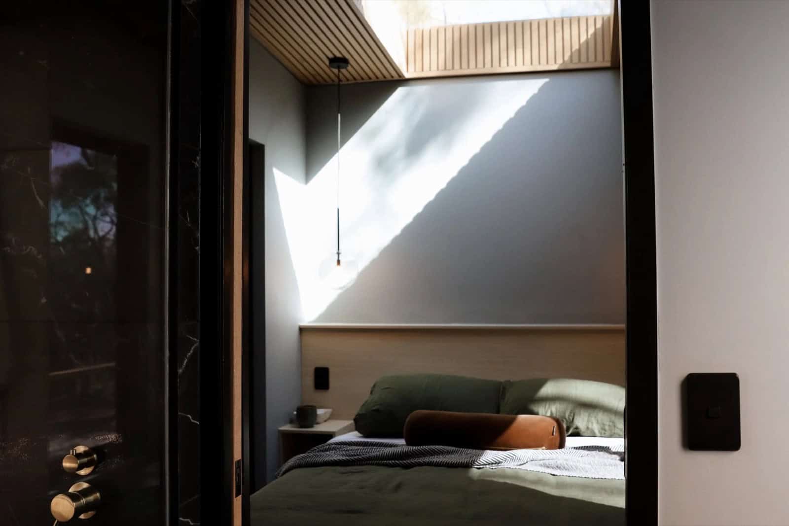 The SHAK bedroom with a queen bed in olive-green linen beneath a large skylight and a timber-slat ceiling, with a glass pendant light